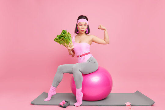 Sporty self confident Asian lady keeps to healthy diet shows muscles raises arms sits on inflated fitness ball does exercises at yoga mat being in good physical shape isolated over pink background