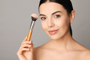 Beautiful young woman applying face powder with brush on grey background, closeup