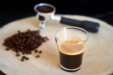 
Espresso in crystal glass with coffee beans and filter holder with out of focus ground coffee on natural wood log