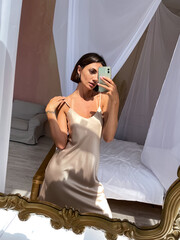 Fit tanned woman in romantic beige silk dress at home take photo selfie on phone in mirror for social media, stories, vertical