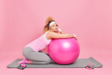 Displeased bored woman leans at fitness ball rests after exercise session dressed in activewear...