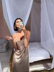 Obraz premium Fit tanned woman in romantic beige silk dress at home take photo selfie on phone in mirror for social media, stories, vertical