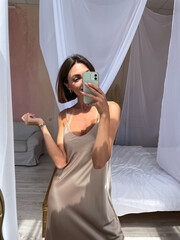 Obraz premium Fit tanned woman in romantic beige silk dress at home take photo selfie on phone in mirror for social media, stories, vertical