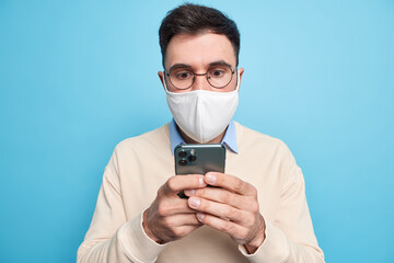 Young brunet man focused surprised at cellular display wears round spectacles checks newsfeed via smartphone dressed casually isolated over blue background. Technology and quarantine concept