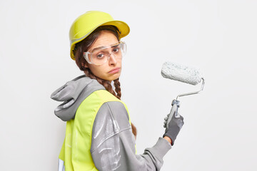 House remodeling and redecoration concept. Sad tired female builder holds paint roller uses construction tool for painting walls wears helmet safety glasses and working clothes isolated on white