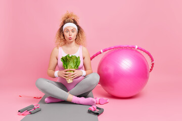 Fototapeta na wymiar Photo of surprised curly woman holds green vegetable has healthy nutrition and active lifestyle sits crossed legs on fitness mat exercises at home isolated on pink wall. Sport dieting wellness concept