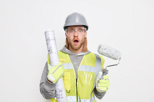 Home improvement concept. Stunned bearded male architect holds blueprint and painting roller wears protective clothes comes on construction site finds out how much work he should do. Shocked laborer