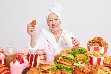 Unhealthy overeating lifestyle. Pleased old lady smiles positively drinks soda eats fast food suffers from gluttony bad eating habits has happy mood isolated over white background wears bathrobe