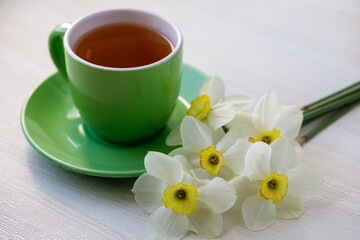 Bouquet white and yellow daffodils and green cup of tea on light wooden table. Close-up, horizontal, top side view