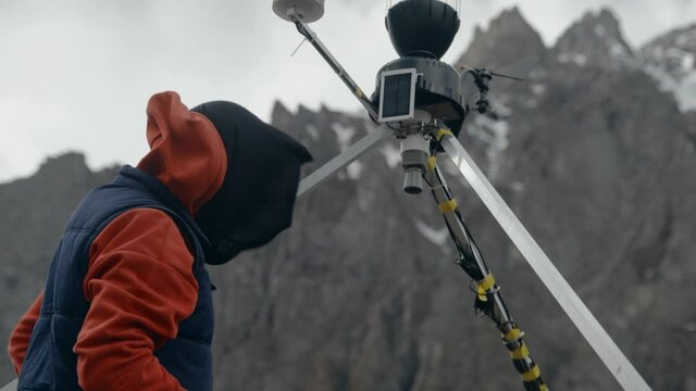 man in glasses in a red hoodie and a blue vest in the mountains checks the operation of the weather station. Mobile weather station on a solar battery. measurement of meteorological conditions