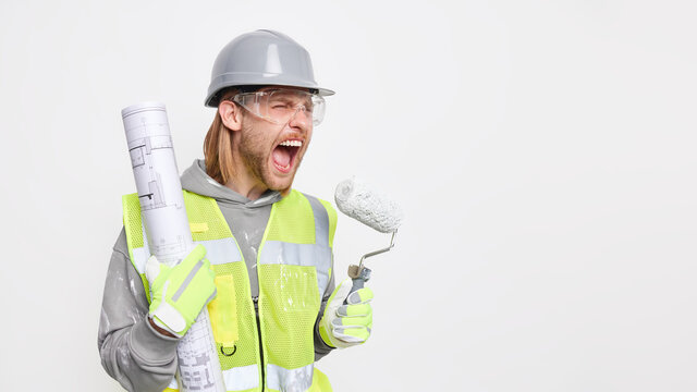 Horizontal shot of irritated male construction worker exclaims negatively holds blueprint and painting roller busy doing home improvement works on architectural plan isolated over white background