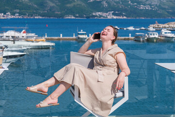 Young pretty European woman on vacations resting and having fun in a cafe terrace with sea view, talking on her smartphone and laughing. Humor, having fun on vacations concept