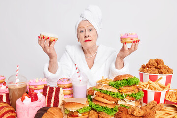 Serious grandmother with wrinked skin dressed in domestic clothes holds two glazed donuts has food addiction eats fast food surrounded by variety of cheat meal isolated over white background