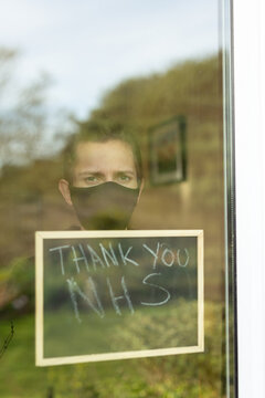 A Young Man With A Face Mask On Looking Out The Window During His Self-isolation At Home, He Is Holding Up A Sign With Thank You NHS Written On It