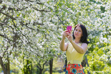 a young girl in a bright yellow-red summer dress takes pictures of herself on a smartphone against the background of a blooming apple orchard