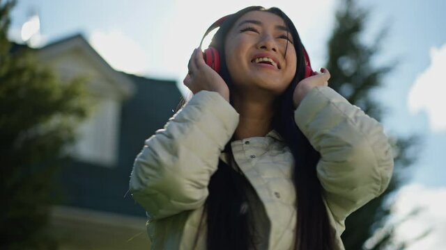 Joyful cheerful Asian young woman in headphones dancing in sunlight spring summer garden outdoors. Portrait of happy carefree relaxed beautiful slim millennial having fun on backyard enjoying hobby