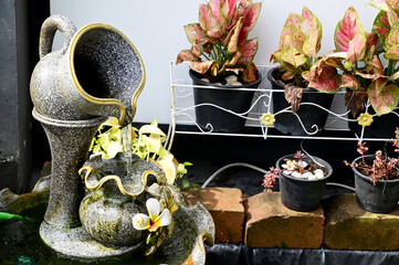 Closeup of A ceramic stucco Jar with water flowing into the fish pond with plants in black pots for decorating the garden to be beautiful in Thailand.