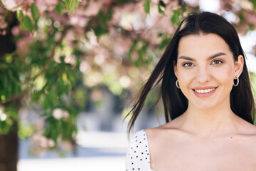 Beautiful smiling confident young ethnic woman pretty face looking at camera posing alone at park on a background of sakura trees. Happy millennial ethnicity girl student professional front portrait.