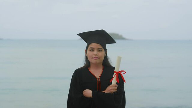 An Asian woman wearing a bachelor's suit with a graduation certificate standing with her arms folded over the sea and sky background.