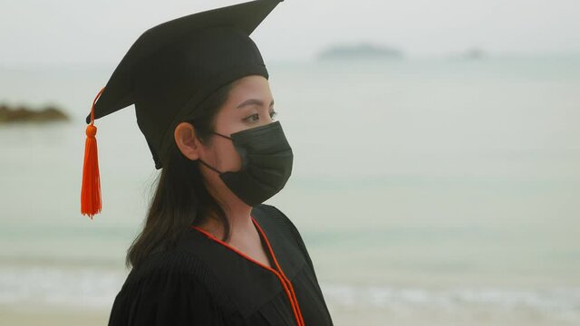 An Asian female graduate who graduated from the university. She walks on a beach with water and waves in the background, wearing a surgical mask to prevent the coronavirus outbreak.