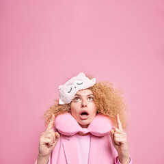 Impressed curly young woman keeps jaw dropped indicates overhead notices something amazing focused above with shocked expression wears sleepmask and neck pillow isolated on pink studio wall.
