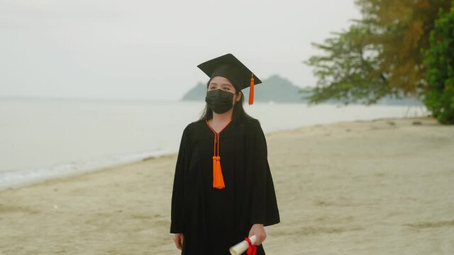 Get your degree during the coronavirus outbreak. An Asian woman wearing a bachelor's suit with a graduation certificate standing with her arms folded over the sea and sky background.
