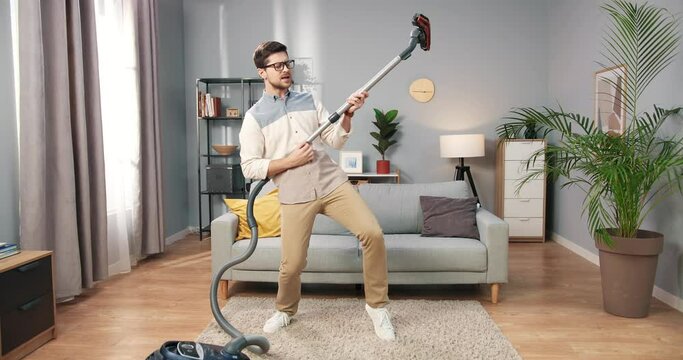 Young stylish Caucasian man with glasses is having fun at home doing daily routine of cleaning living room using vacuum cleaner as electric guitar.