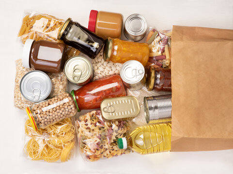 Food donations such as pasta, rice, oil, peanut butter, canned food, jam and in a paper bag on white wooden table 