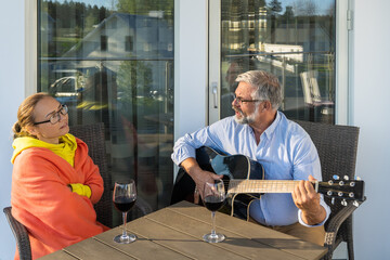 Couple man and woman relaxing outside the house on terrace or balcony. Mid aged female and senior male smiling enjoying red wine in glasses. Bearded man imposing musician plays the guitar and sings.