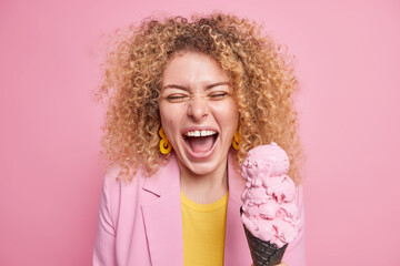 Joyful pretty woman has fun laughs happily holds tasty ice cream expresses positive sincere emotions dressed elegantly poses against pink background. Positive female model with appetizing gelato