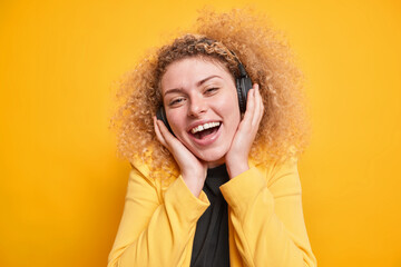 Portrait of happy curly woman keeps hands on stereo wireless headphones smiles broadly being in good mood dressed in formal jacket enjoys spare time isolated over yellow background. Positive emotions