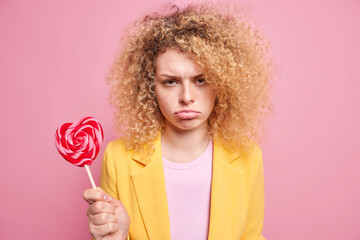 Sad frustrated curly haired woman has sweet tooth holds candy on stick cannot afford eating it has...