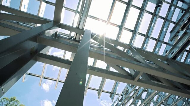 Bright sun shining through steel metallic building framing structure, low angle perspective on construction site with light flare.