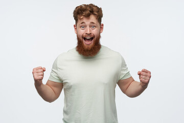 studio shot of young redhead bearded male wears blank t-shirt, raise his fist and yelling while watching football match. victory concept. isolated over white background