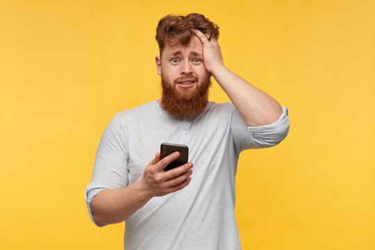 amazed young man with a big red beard staring into camera with a shocked facial expression, holds his phone, get some good news. isolated over yellow background.