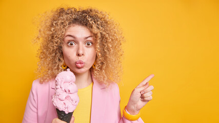 Lovely surprised woman with curl bushy hair folds lips indicates away on blank space holds tasty ice cream invites to visit some place and taste delicious dessert isolated over yellow background