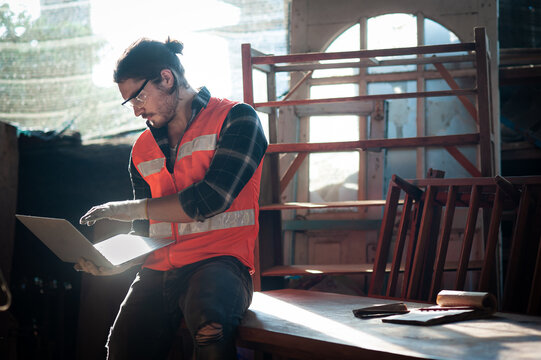 Young and handsome caucasian carpenter in the carpentry shop is working and checking blueprint in laptop That customers send by email to make furniture according to orders and repair a furniture.