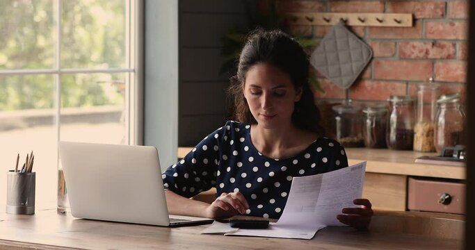 Pleasant young hispanic woman sitting at table with computer, calculating paper bills, managing bank loan mortgage payments, planning monthly budget, paying taxes, satisfied with enough money.