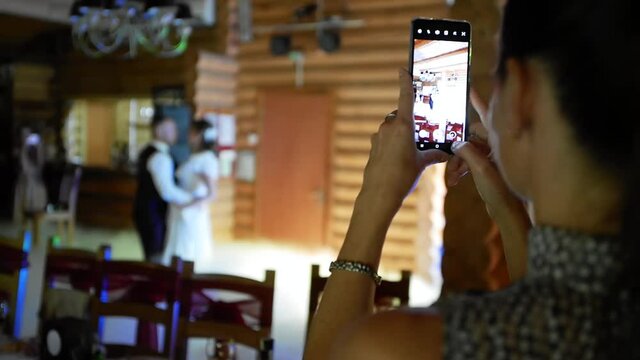 A woman photographs a couple of newlyweds during a wedding dance on a cell phone. A young girl shoots a video on a smartphone during a wedding while holding a gadget in her hands.