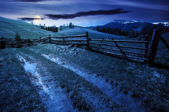 mountainous rural landscape at night in spring. path through grassy field. wooden fence on rolling hills. snow capped ridge in the distance. wonderful countryside scenery in full moon light