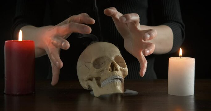 Hand with magician skull. A woman makes magic with a human skull and burning candles on the table in the dark in the room.