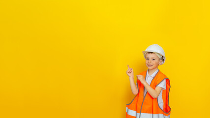 Portrait of a smiling boy in a white hard hat and orange signal vest, pointing with his finger at a free space for text. Yellow background.