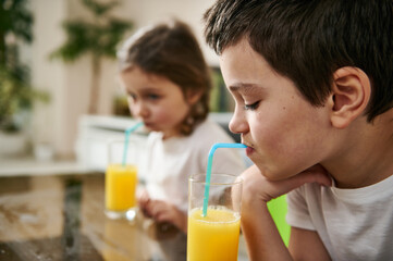 Little boy drinking orange juice, sitting at the table next to his sister blurred on the background