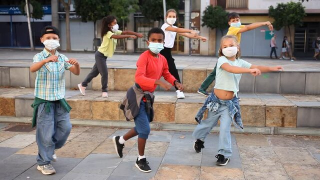 Positive girls and boys in face masks training hip hop on city street, outdoor dance class for kids at pandemic situation