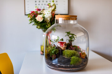 Terrarium with plants - ginseng ficus, miniature house, miniature world. A forest in glass in the company of cut flowers on the table in front of a Scandinavian poster. Minimalist interio