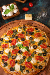 In the photo we see pizza on a wooden stand. The pizza is beautifully decorated with tomatoes, olives, cheese, green mint leaves. Close-up. Macro photography. Careful viewing.