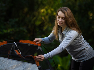 In the photo, a young smiling woman is resting in nature. The woman lays out the tent. Evening time. In the background is a photo of a green forest. Close-up. There is a place for your insert.