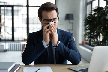 Frustrated businessman, office employee, manager blowing running nose, covering face with paper tissue at workplace in office. Man feeling unwell, suffering from flu infection, spring allergy.