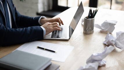 Businessman using laptop among crumpled paper drafts messy in office. Professional typing important document on keyboard, writing article or report, working at computer. Close up of hands