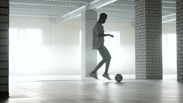 Young black guy trains with a soccer ball at indoors. Bright sunlight at background